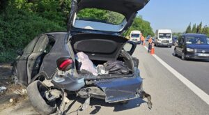 Otoyolda hasarlı otomobilin açık bagajı ve önünde yol kenarı ile trafik görevlileri, kaza sonrası araçlar görünüyor.