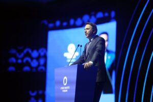 Koyu takım elbiseli bir konuşmacı Zero Waste Forum yazılı kürsüde mikrofonlar önünde konuşuyor.