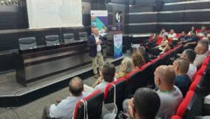 Konferans salonunda konuşan bir kişi, önde kırmızı koltuklarda oturan dinleyiciler ve yanlarında AB hibe pankartı görünüyor.