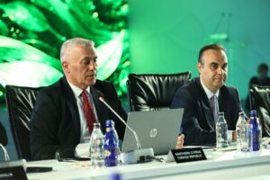 Yeşil ışıklı sahnede iki erkek takım elbiseli, masada dizüstü bilgisayar ve mikrofonlarla toplantı yapıyor