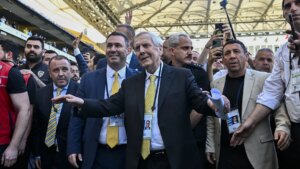 Stadyumda kalabalık içinde iki yönetici, biri elinde telefonuyla fotoğraf çektiriyor, diğeri elini kaldırmış durumda