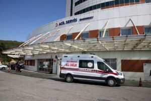 Acil servis tabelalı hastane binası önünde kırmızı şeritli ambulans park halinde.