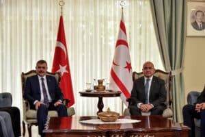 Başörtü oturma odasında iki erkek, Türk bayrakları önünde koltuklarda oturuyor ve masa üzerinde çaydanlık bulunuyor.