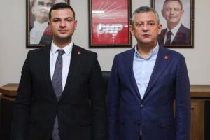 Takım elbiseli iki kişi, bir oda içinde duvarda asılı portrelerin önünde yan yana poz veriyor.