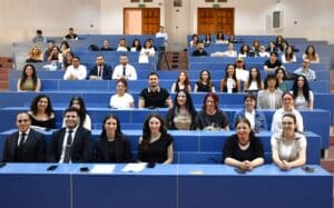 Sınıf sıralarında toplanmış, kürsü önünde poz veren katılımcılar, üniversite salonunda grup fotoğrafı çekimi yapıyor