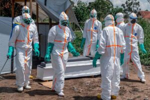 Beyaz tulumlu sağlık personeli, eldiven ve yüz maskeleriyle dış ortamda Ebola benzeri koruma ekipleriyle yürürken görülüyor.