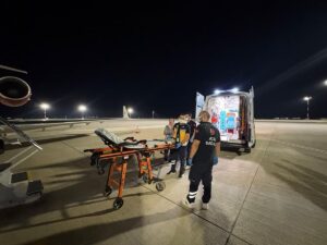 Gece havalimanında ambulansın yanında sedyeyi taşıyan sağlık ekipleri ve uçak rampası görünüyor.