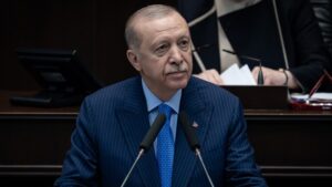 Parlamento kürsüsünde iki mikrofon önünde konuşan mavi takım elbiseli Recep Tayyip Erdoğan portresi