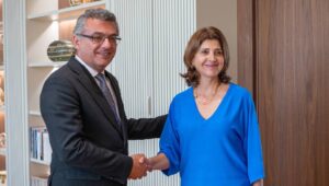 Takım elbiseli bir erkek ve mavi bluzlu kadın ofiste tokalaşırken gülümsüyor.