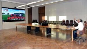 Toplantı salonunda uzun masa üzerinde oturan kişi, karşıda konferans ekranı ve arka sırada sandalyeler bulunuyor.