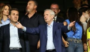 Siyah takım elbiseli ve mavi ceketli bir grup içinde, kollarını iki yana açmış beyaz saçlı bir adam konuşur gibi görünüyor.