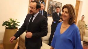 Siyah takım elbiseli bir erkek ile mavi elbiseli bir kadın, resmi bir ortamda gülümseyerek yan yana duruyor.