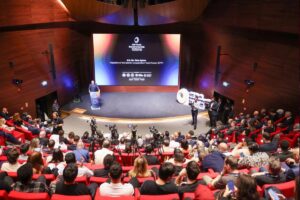 Konferansta sahnede konuşmacı kürsüde sunum yaparken seyirci kırmızı koltuklarda ve kameralar toplantıyı görüntülüyor
