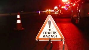 Kırmızı gece yolunda Trafik Kazası uyarı işareti ve şerit üzerinde ışıklı araçlar görünüyor.