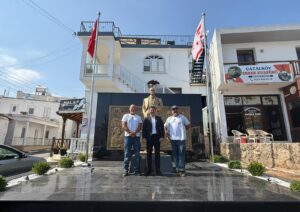 Atatürk büstünün önünde üç kişinin durduğu, Türk bayraklı ve tabelalı dükkân binası.