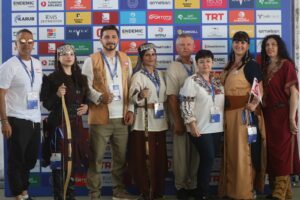 Okçuluk yarışması ödül töreninde takım halinde duran, mavi madalyalı sekiz kişi ve arka planda sponsor panosu