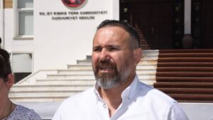 Beyaz gömlekli sakallı bir adam, binanın önünde kameraya doğru konuşurken görülüyor.