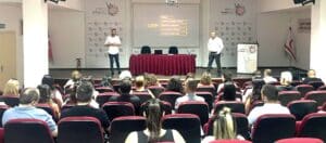 Seminer salonunda konuşmacılar kürsüde eğitim verirken izleyiciler kırmızı koltuklarda oturuyor