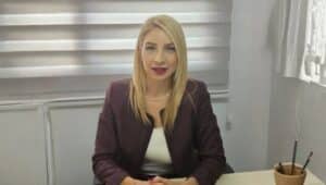 Masa başında konuşan sarı saçlı kadın, mor ceket ve beyaz bluz giyiyor.