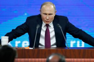 Koyu takım elbiseli Vladimir Putin, masada iki mikrofonla konuşurken kollarını açmış halde oturuyor.