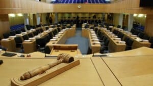Bir meclis salonunda sıralar halinde oturma düzeni ve masalarda mikrofonlar ile kürsü görülüyor.