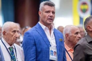 Mavi blazerli, yaka kartlı bir adam konuşurken yanında başka yaşlı erkeklerin bulunduğu kalabalık etkinlik fotoğrafı