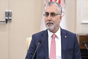 Gözlüklü gri sakallı bir adam, pembe kravatlı takım elbise ile kürsüde mikrofonlara konuşuyor.
