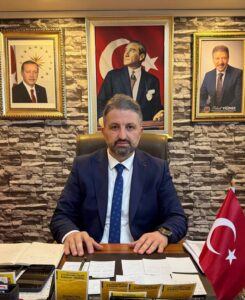Türk bayrağı yanında masada oturan takım elbiseli bir adam; arka planda çerçeveli Atatürk portreleri bulunan ofis ortamı