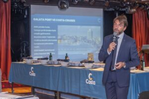 Galata limanı ve Costa kruvaziyerleri hakkında sunum yapan, elinde mikrofon bulunan takım elbiseli konuşmacı.