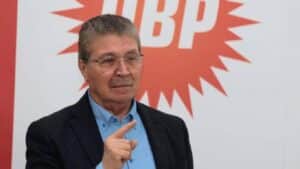 Gözlüklü bir erkek, kırmızı-beyaz arka planda konuşuyor ve elini işaret ederken poz veriyor.