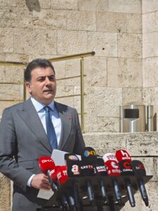 Mikrofon yığınları önünde konuşan takım elbiseli bir adam, taş duvarın yanında basın açıklaması yapıyor