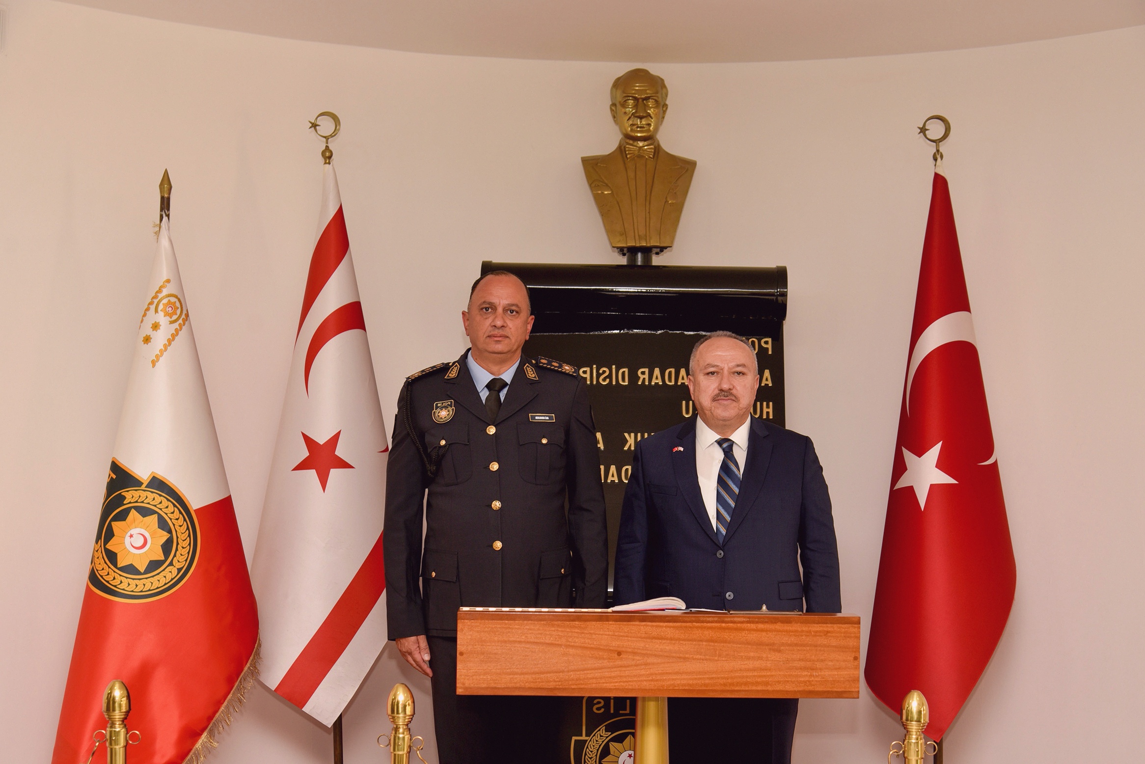 Türk bayrakları önünde biri üniformalı iki erkek, büst heykeli ve yazı masası yanında poz veriyor.