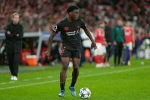 Siyah formayla sahada topun yanında duran futbolcu, tribünlerde yedekler ve antrenör görünür.