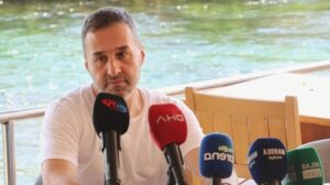 Su kenarında teknede oturan erkek gazeteci, üzerinde mikrofonlarla kameraya konuşurken görülüyor.