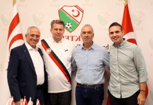 Dört erkek, Türkiye ve bayraklarla süslü federasyon duvarında, futbol forması ve logo önünde birlikte poz veriyor.