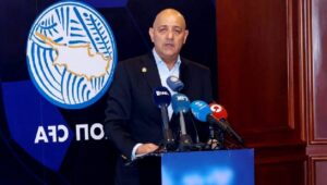 Mikrofonlar önünde konuşan takım elbiseli bir adam, arkasında Rum Futbol Federasyonu logosu ve mavi zemin.
