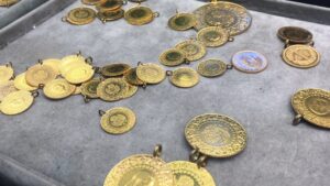 Gri kadife üzerinde altın renkli madeni para ve kolye parçaları serili halde görünüyor.