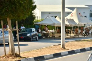 Çadırlarla kurulan yardım alanında masalar ve sandalyeler, yanında park etmiş araçlar görünüyor.
