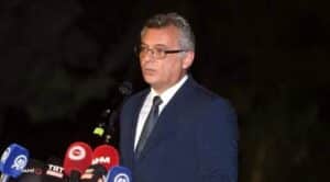 Siyah zemin önünde konuşan gözlüklü takım elbiseli bir erkek, mikrofonlar ve basın açıklaması panelleri önünde duruyor.