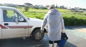 Önlük giymiş bir kişi, Güney Kıbrıs’ta yol kenarında beyaz bir arabanın yanında kapıdan bir şey aldı.