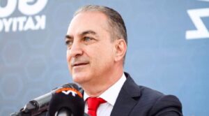Mikrofonlar önünde siyah takım elbiseli, kırmızı kravatlı bir adam konuşurken görülüyor.