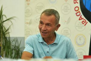 Arka planda KOD 12 ve benzeri yazıların olduğu basın toplantısı fonu önünde mavi polo gömlekli bir adam gülümserken oturuyor