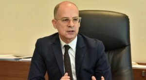 Siyah takım elbiseli, gözlüklü bir erkek siyah koltukta otururken konuşuyor.