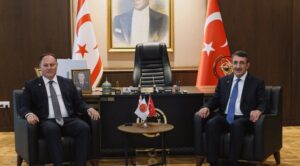 Türkiye ve siyah bayraklı toplantı salonunda iki kişi koltuklarda oturuyor, arada küçük masa ve Türk bayrakları var