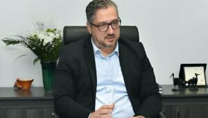 Siyah takım elbiseli, gözlüklü bir adam ofiste koltuğunda otururken konuşuyor, arka planda çiçekler ve çerçeve var.