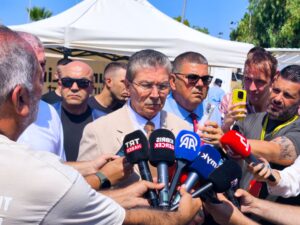 Süt beyaz ceketli bir adam, arka plandaki gazeteciler ve mikrofonlarla çadır altında açıklama yapıyor.