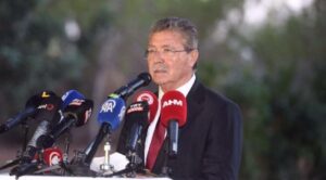 Mikrofonlar önünde konuşan takım elbiseli bir erkek, beyaz masa başında görüntüleniyor.