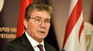 Gözlüklü yaşlı bir erkek, yanında ABD ve Türk bayrakları varken konuşma sırasında mikrofonla konuşuyor.