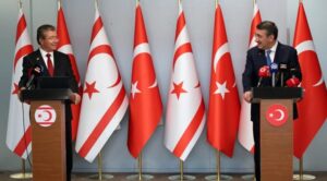 Kürsüde konuşan iki siyasetçi, arka planda Türkiye ve KKTC bayrakları bulunan kapalı ortamda basın toplantısı.
